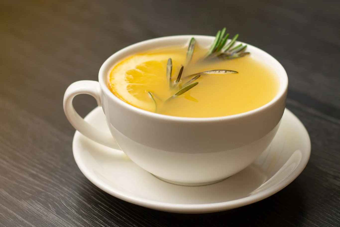 Ze względu na wyjątkowy smak i aromat rozmaryn dodaje się do wielu potraw, a nawet napojów i deserów Biała filiżanka wypełniona żółtym napojem ozdobionym plasterkiem cytryny oraz gałązkami rozmarynu, ustawiona na spodku na ciemnym blacie.