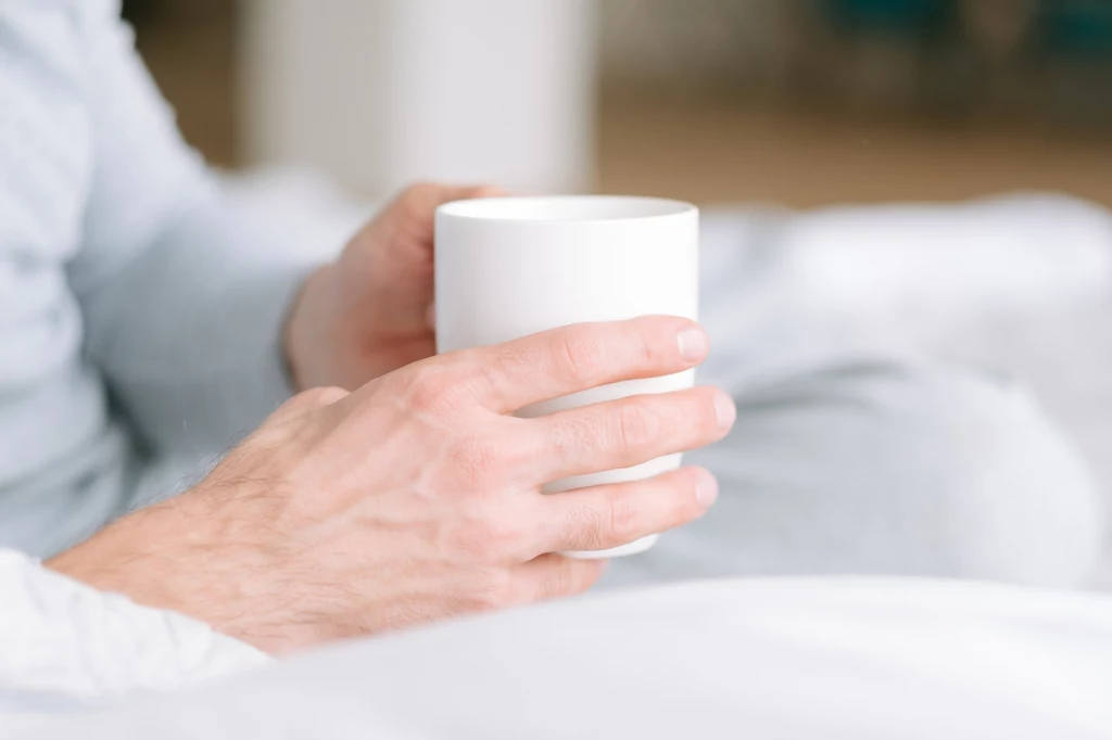 Naturalny sposób na dobry sen znajdziesz wśród ziół Dłonie osoby trzymające biały kubek w lekko rozmytym, jasnym otoczeniu sugerującym domowe lub relaksujące warunki.