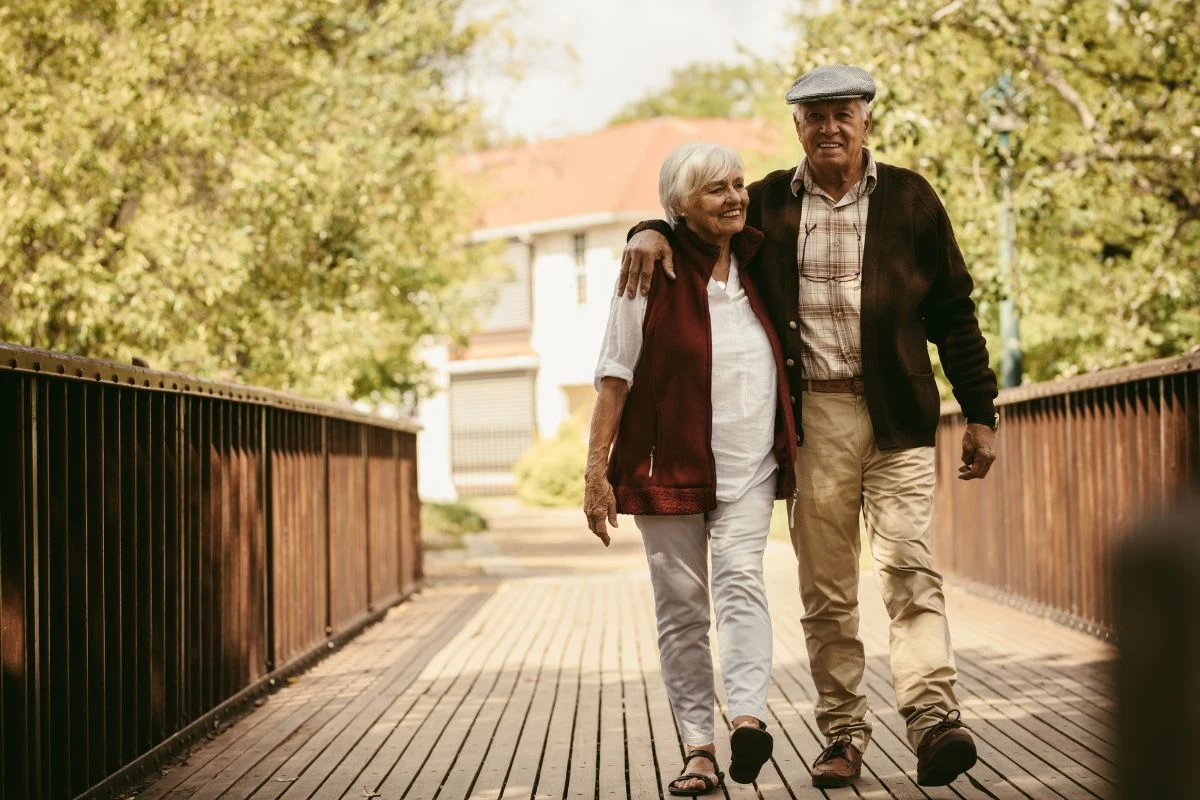 Starsza para obejmująca się podczas spaceru po drewnianym moście otoczonym zielenią, oboje wyglądają na zrelaksowanych i szczęśliwych, ubrani w wygodne ubrania, w tle widoczny rozmazany budynek mieszkalny.