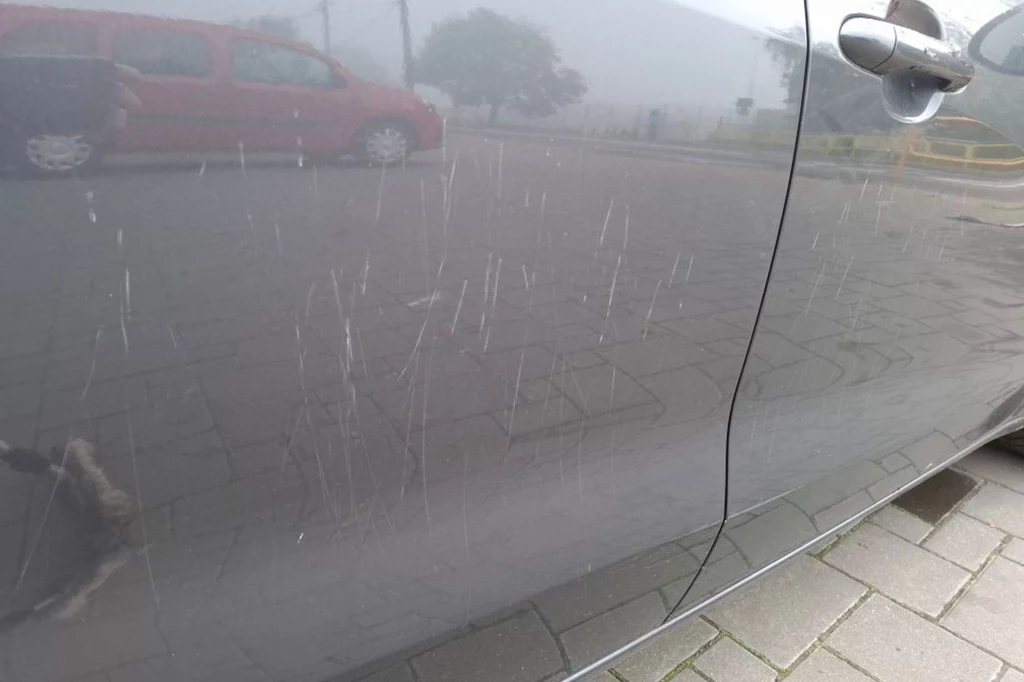 Głębokie rysy na karoserii samochodu. Mieszkaniec Piątnicy pokazuje straty Boczne drzwi samochodu w ciemnym kolorze z licznymi długimi rysami widocznymi na lakierze, w tle zaparkowany czerwony samochód oraz brukowany chodnik.