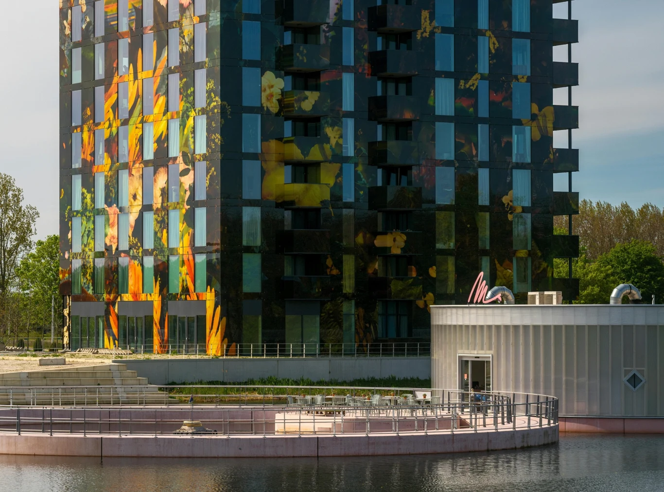 Fot. pexels.com/ Haberdoedas Photography (zdjęcie poglądowe) Nowoczesny, wielopiętrowy budynek z kolorową, kwiatową grafiką na szklanej elewacji, przed którym znajduje się niewielki, okrągły budynek oraz sztuczny zbiornik wodny, otoczony zielenią.