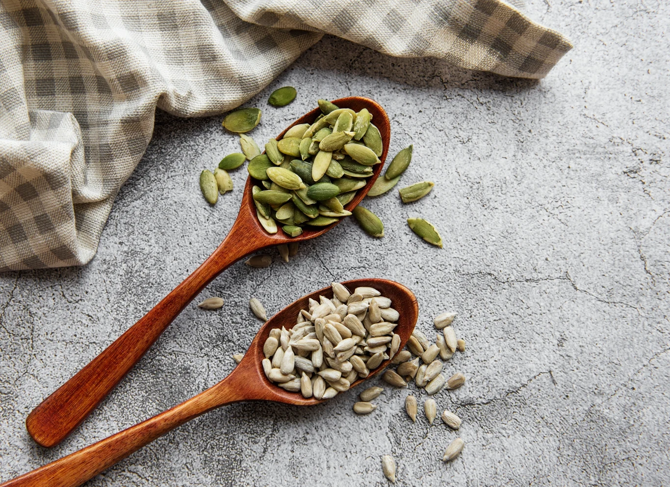 Dwie drewniane łyżki z nasionami słonecznika i pestkami dyni leżące na marmurowej powierzchni obok beżowo-szarej ściereczki.