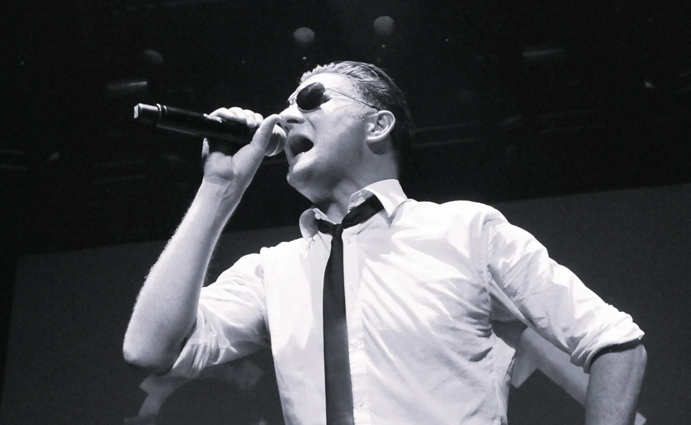 Nie żyje Douglas McCarthy Mężczyzna w okularach przeciwsłonecznych śpiewający do mikrofonu na scenie, ubrany w białą koszulę i czarny krawat, dynamiczne oświetlenie koncertowe w tle.