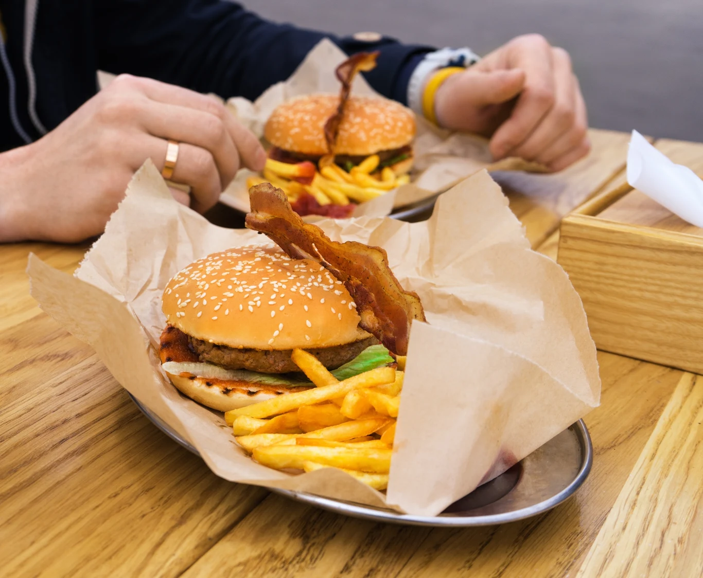 Soczysty hamburger z bekonem oraz porcją złocistych frytek leżą na metalowej tacy wyłożonej papierem, w tle widoczna osoba trzymająca frytki.