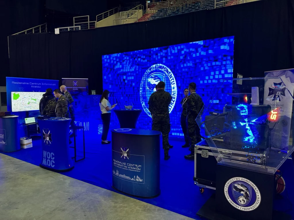 CYBERSEC EXPO & FORUM 2025. Tak zaprezentowało się Regionalne Centrum Informatyki Kraków Wojsko w mundurach rozmawia i zapoznaje się z multimedialną ekspozycją w centrum prezentacyjnym ze ścianą ekranów LED, stoiskami informacyjnymi oraz sprzętem wojskowym, całość utrzymana w niebieskiej kolorystyce i otoczeniu technologicznym.