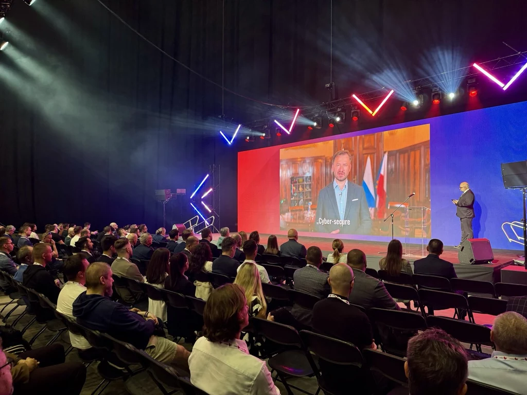 Prelekcje na scenie CYBERSEC EXPO & FORUM 2025 Sala konferencyjna z dużą liczbą uczestników siedzących w rzędach, scena oświetlona kolorowymi światłami LED, na telebimie widoczny mężczyzna przemawiający w eleganckim gabinecie z flagą w tle, osoba prowadząca na scenie.