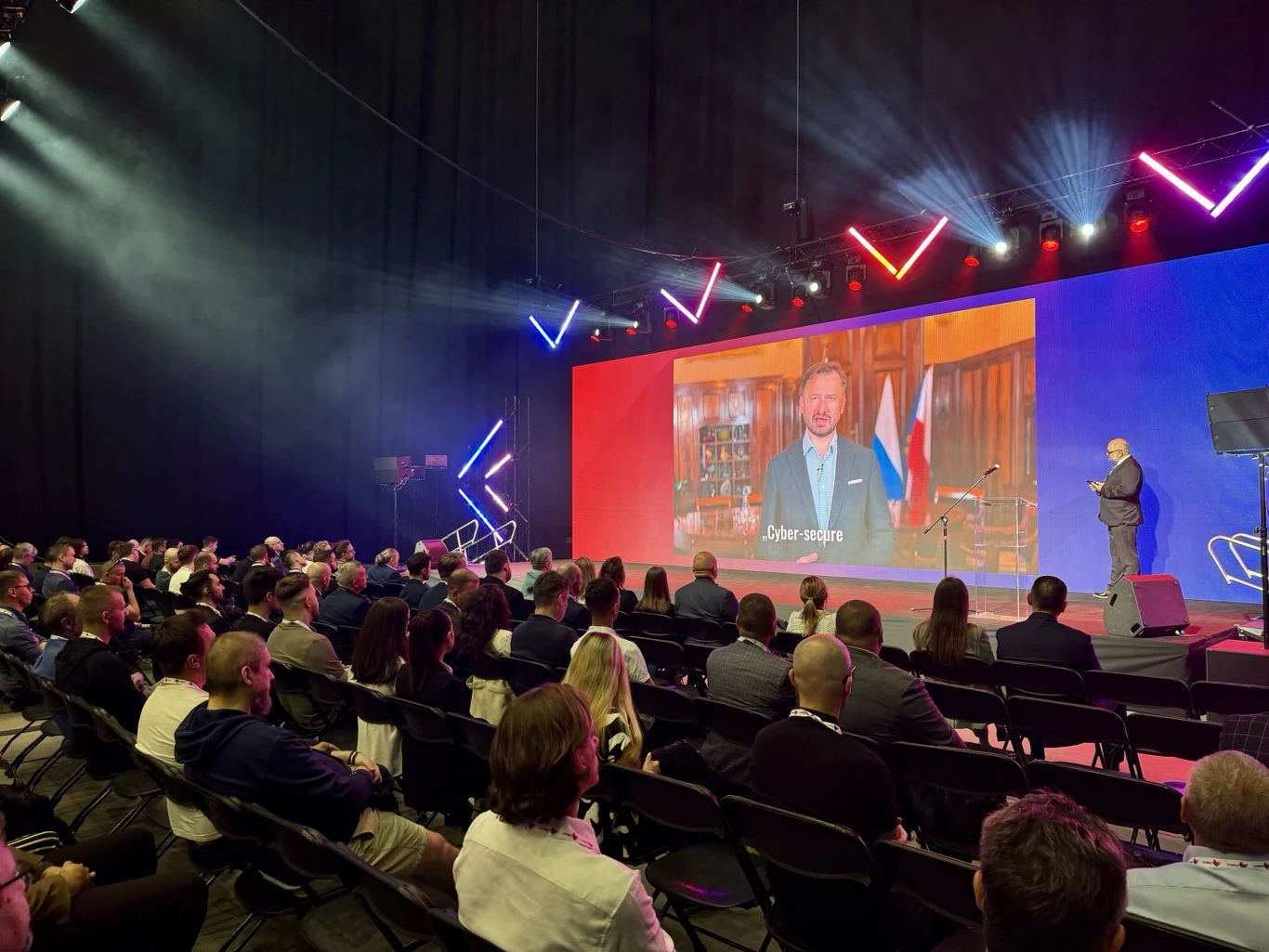 Prelekcje na scenie CYBERSEC EXPO & FORUM 2025 Sala konferencyjna z dużą liczbą uczestników siedzących w rzędach, scena oświetlona kolorowymi światłami LED, na telebimie widoczny mężczyzna przemawiający w eleganckim gabinecie z flagą w tle, osoba prowadząca na scenie.
