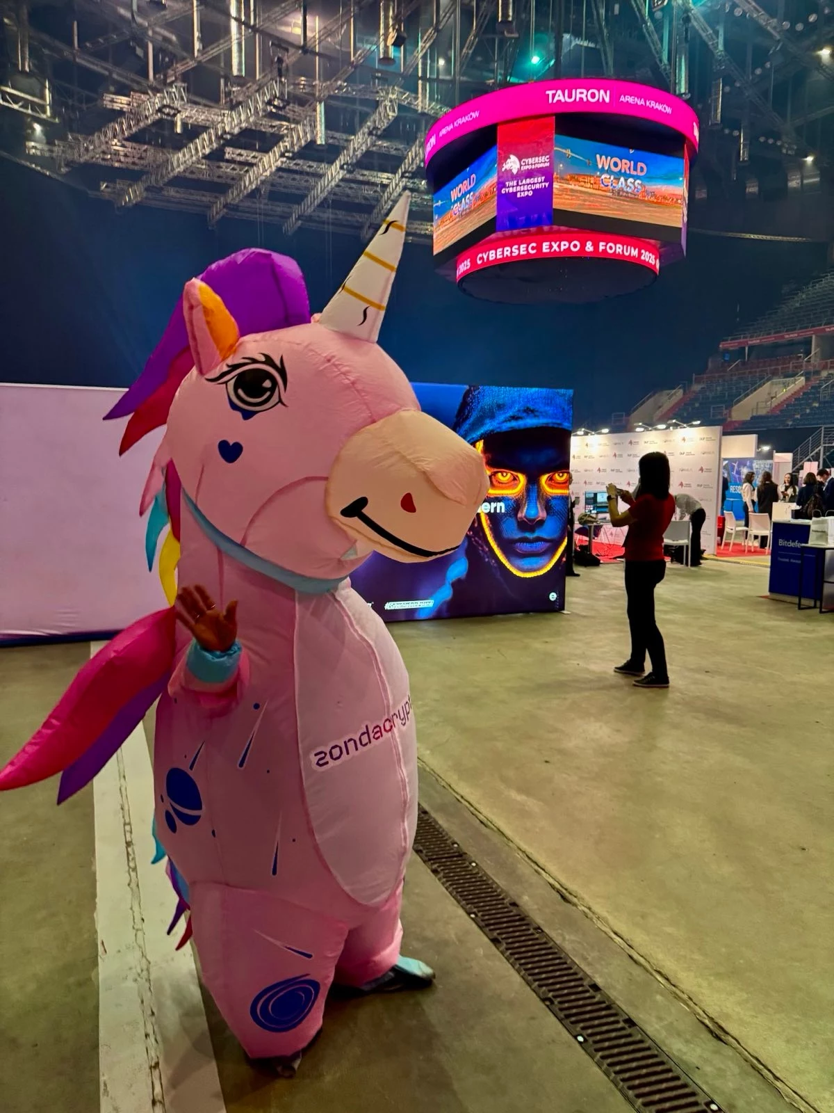 Jak na konwencie. Na targach CYBERSEC EXPO & FORUM 2025 nie zabrakło też elementów humorystycznych. Do zobaczenia za rok! Kolorowy kostium jednorożca z logo firmy, stojący wewnątrz nowoczesnej hali targowej, w tle duży ekran LED z grafiką twarzy i pojedyncze sylwetki ludzi oraz główna scena wydarzenia.