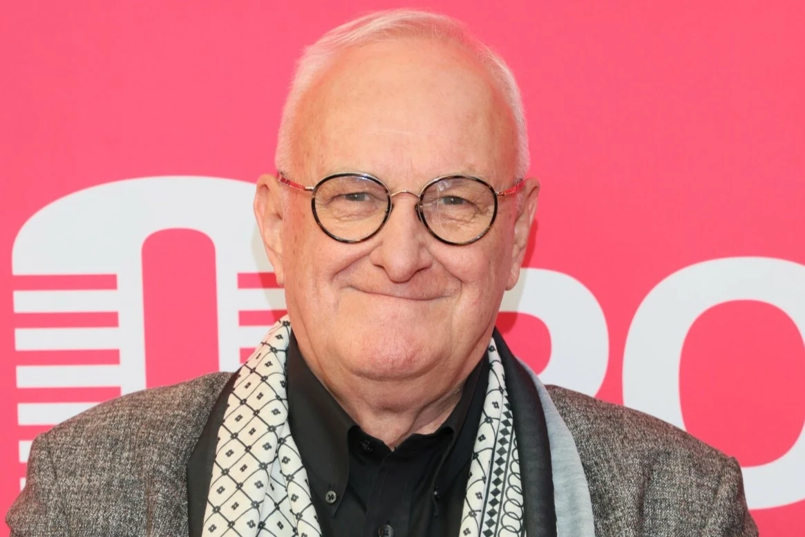 Jacek Cygan to legenda polskiej muzyki rozrywkowej Starszy mężczyzna w okularach i szaro-czarnej marynarce, uśmiechnięty, stoi na tle grafiki z różowym napisem i białymi literami.