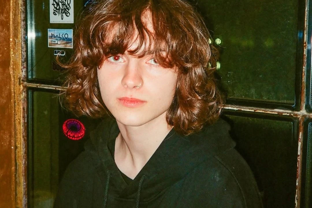 Kosma Król Młoda osoba z dłuższymi, brązowymi włosami i jasną cerą, ubrana w czarną bluzę, stoi przed szybą z naklejkami, patrząc prosto w obiektyw.