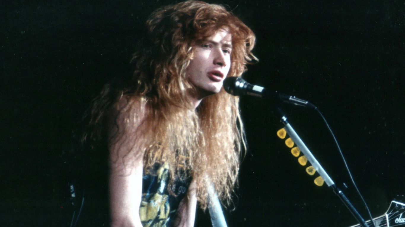 Dave Mustaine (1988) Młody mężczyzna o długich rudych włosach śpiewa do mikrofonu, trzymając gitarę elektryczną podczas koncertu na scenie, w tle ciemność oraz fragment statywu z rzędem żółtych kostek do gitary.