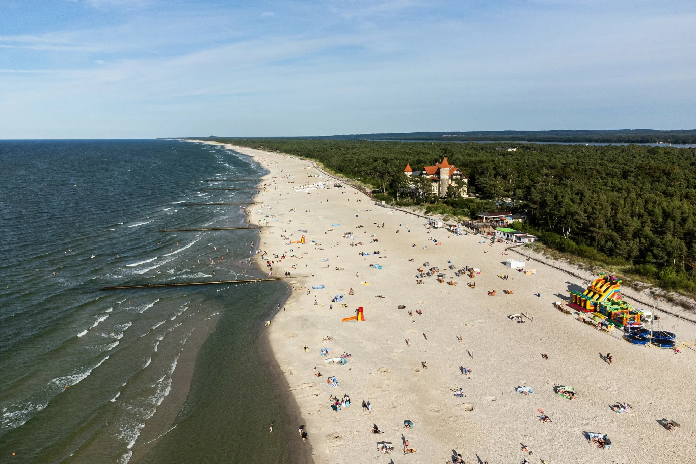 Szeroka piaszczysta plaża nad morzem z licznymi turystami, w tle pas lasu i budynek z wieżyczkami, na prawo kolorowe atrakcje dla dzieci.