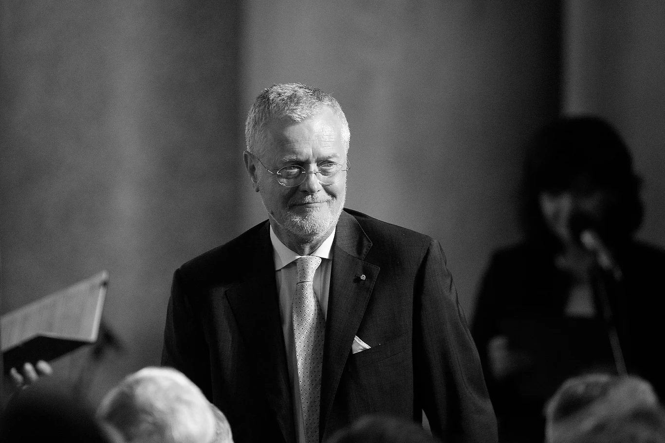 Piotr Nowina-Konopka nie żyje Starszy mężczyzna w garniturze stoi uśmiechnięty w formalnym otoczeniu, na tle rozmytej postaci i mikrofonu, sugerujących oficjalne wydarzenie lub uroczystość.