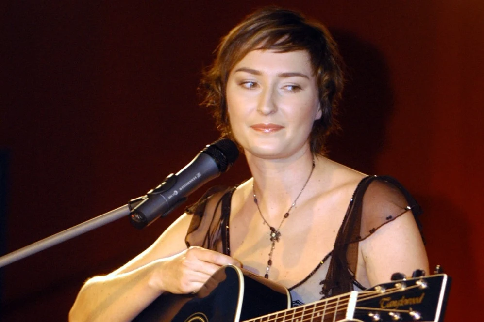 Młoda kobieta z krótkimi brązowymi włosami trzyma czarną gitarę akustyczną i stoi przy mikrofonie, ubrana w elegancką, ciemną sukienkę z naszyjnikiem, z delikatnym, neutralnym wyrazem twarzy na tle rozmytego, ciepłego tła.