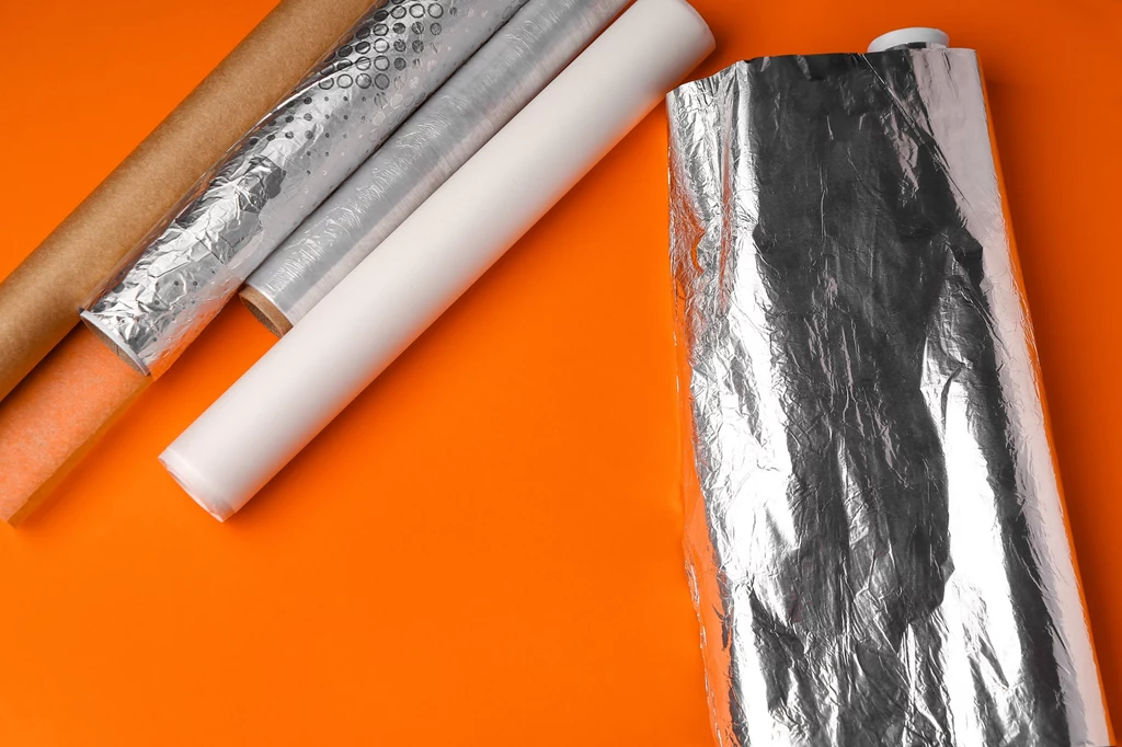 Końcówki banana owijamy folią aluminiową lub spożywczą przezroczystą, aby ograniczyć wydzielanie etylenu Rulony folii aluminiowej, papieru do pieczenia, folii bąbelkowej oraz papieru pakowego ułożone na pomarańczowym tle, wyraźnie widoczne tekstury materiałów.