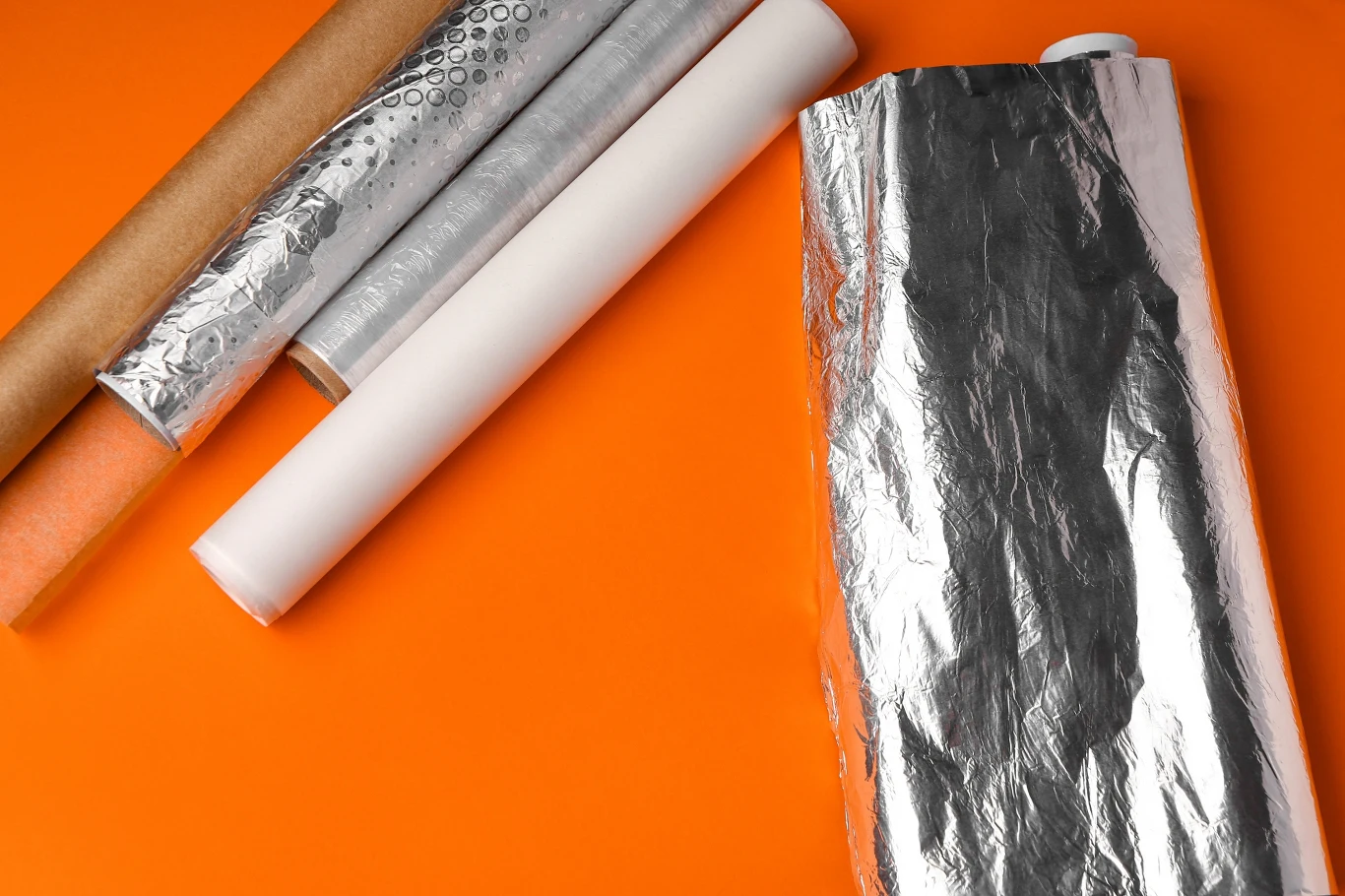 Rulony folii aluminiowej, papieru do pieczenia, folii bąbelkowej oraz papieru pakowego ułożone na pomarańczowym tle, wyraźnie widoczne tekstury materiałów.