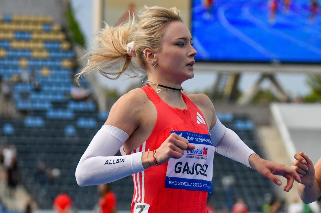 Wiktoria Gajosz Zawodniczka w czerwonym stroju sportowym podczas biegu na stadionie lekkoatletycznym, widoczne kibice na trybunach w tle oraz inny zawodnik po prawej stronie kadru.