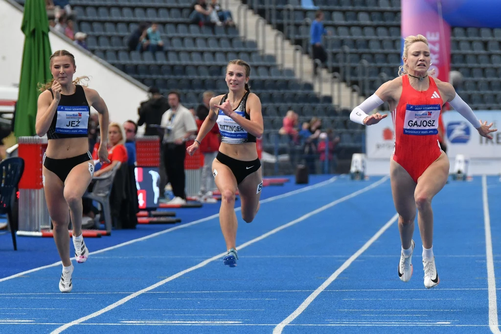 Wiktoria Gajosz (pierwsza z prawej) Trzy zawodniczki biegnące po niebieskim tartanie podczas zawodów lekkoatletycznych, widoczne są linie torów bieżni oraz trybuny w tle, zawodniczka po prawej na finiszu wyprzedza pozostałe
