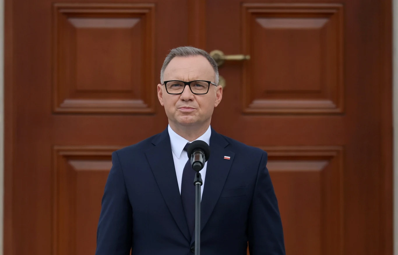 Prezydent Andrzej Duda Mężczyzna w garniturze i okularach stoi przed mikrofonem na tle dużych, drewnianych drzwi, wyrażając powagę sytuacji.