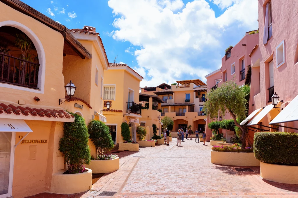 Porto Cervo to jeden z najbardziej znanych kurortów Sardynii Kolorowe, niskie budynki z jasnymi elewacjami wyznaczają wąską, brukowaną uliczkę otoczoną zielonymi roślinami w dużych donicach; na tle nieba z chmurami widoczna jest grupa spacerujących osób.