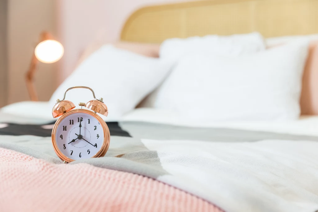 Sleepmaxxing obiecuje bardziej efektywne spanie Złoty budzik z tradycyjną tarczą wskazujący godzinę stoi na pościeli na łóżku, w tle widoczne miękkie poduszki oraz oświetlenie nocnej lampki, całość utrzymana w jasnych oraz pastelowych kolorach