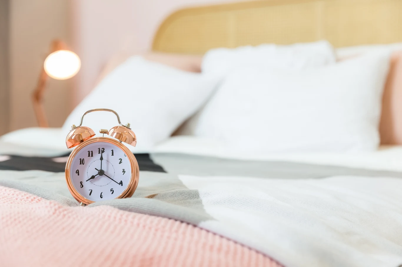 Złoty budzik z tradycyjną tarczą wskazujący godzinę stoi na pościeli na łóżku, w tle widoczne miękkie poduszki oraz oświetlenie nocnej lampki, całość utrzymana w jasnych oraz pastelowych kolorach