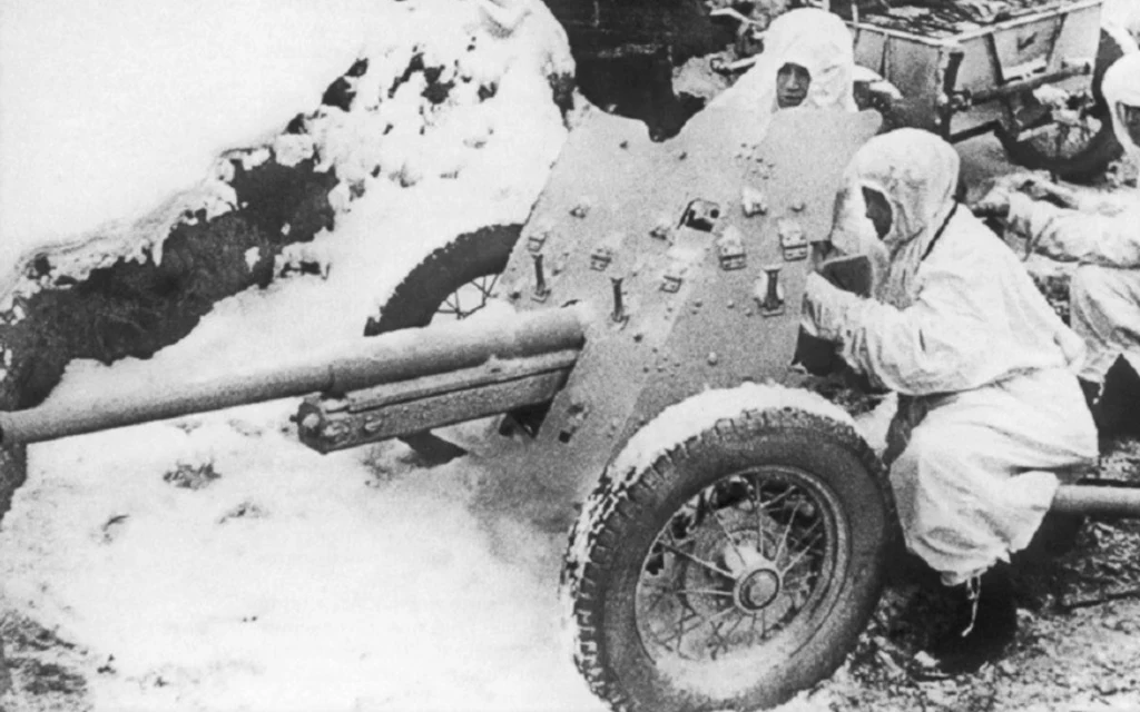 Armata przeciwpancerna 45mm wz. 1937 (53-K) Żołnierze w zimowych białych kombinezonach obsługują armatę przeciwpancerną na zaśnieżonym terenie, wokół widoczne są elementy sprzętu wojskowego.
