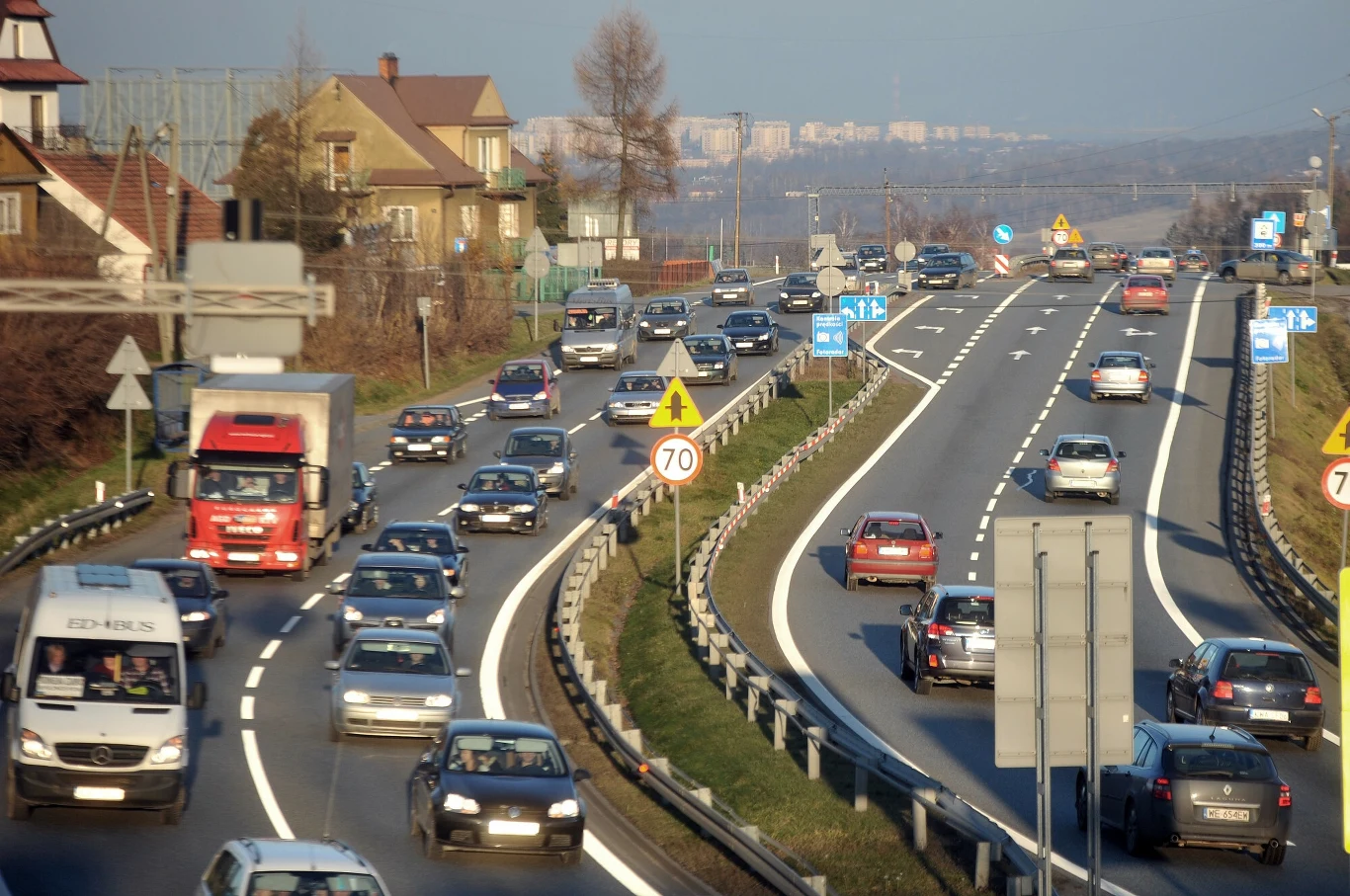 Natężenie ruchu na Zakopiance rośnie z każdym rokiem. Budowa nowej drogi jest niezbędna Ruchliwa droga szybkiego ruchu z wieloma samochodami osobowymi i ciężarówką, barierkami oddzielającymi pasy ruchu, widocznymi znakami drogowymi, budynkami mieszkalnymi po lewej stronie oraz panoramą miasta w oddali.