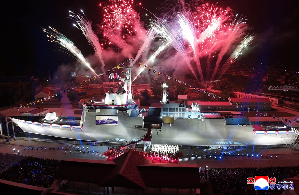 Ceremonia wprowadzenia okrętu Kang Kon do służby Duży okręt wojenny oświetlony podczas nocnej ceremonii, nad statkiem efektowne pokazy fajerwerków, wokół zgromadzenie ludzi i flagi międzynarodowe