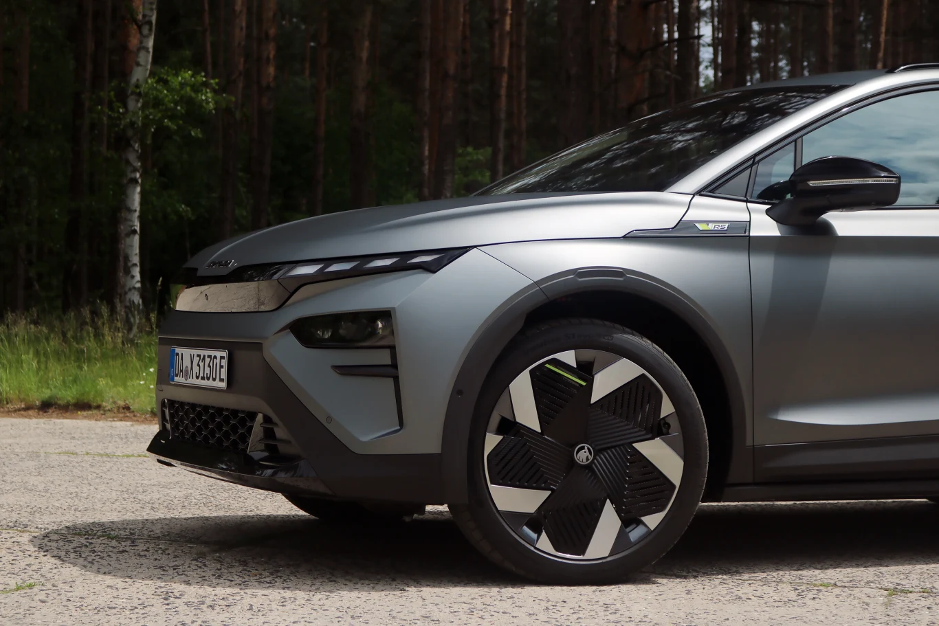 Elroq RS już w standardzie oferuje adaptacyjne zawieszenie DCC. Nowoczesny samochód elektryczny w matowym szarym kolorze, widoczne przednie reflektory, wyraźny wzór na feldze koła oraz fragment tablicy rejestracyjnej, tło stanowi las.
