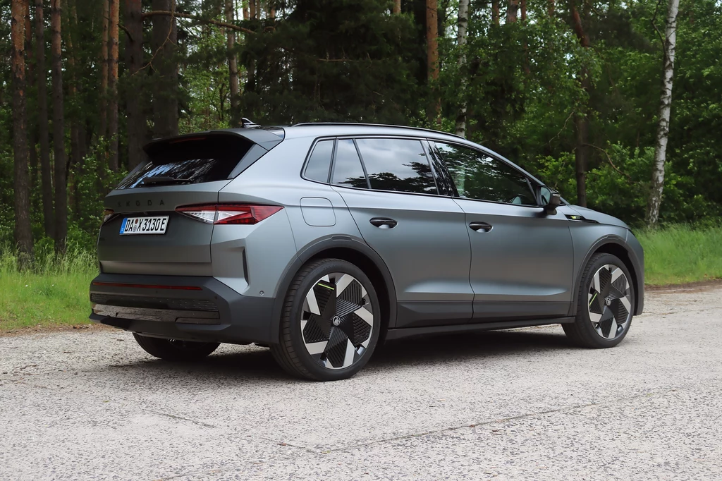 Z tyłu auta uwagę zwraca charakterystyczna dla modeli RS odblaskowa listwa. Szary SUV marki Škoda Enyaq zaparkowany na leśnej drodze asfaltowej, w otoczeniu zielonych drzew i naturalnej roślinności; auto widoczne z tyłu, z zamkniętymi szybami i wyróżniającymi się felgami.