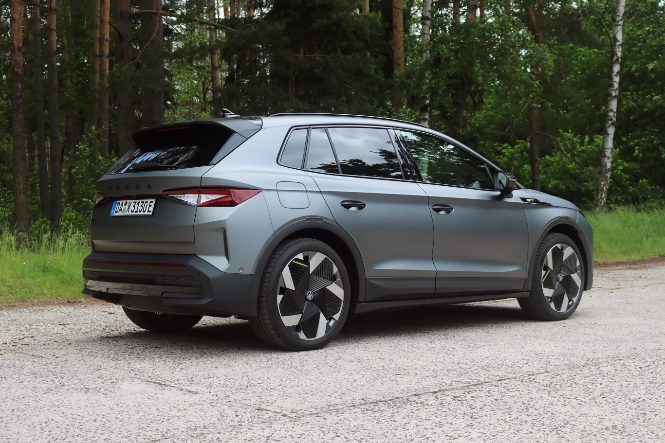 Z tyłu auta uwagę zwraca charakterystyczna dla modeli RS odblaskowa listwa. Szary SUV marki Škoda Enyaq zaparkowany na leśnej drodze asfaltowej, w otoczeniu zielonych drzew i naturalnej roślinności; auto widoczne z tyłu, z zamkniętymi szybami i wyróżniającymi się felgami.