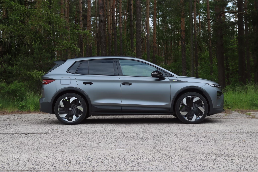 Nowe 21-calowe felgi z aerodynamicznym wykończeniem są zarezerwowane wyłącznie dla modelu RS. Srebrny samochód SUV zaparkowany na tle leśnej drogi, otoczony gęstym lasem iglastym, widoczny z boku, z dużymi alufelgami i czarnymi elementami wykończenia.