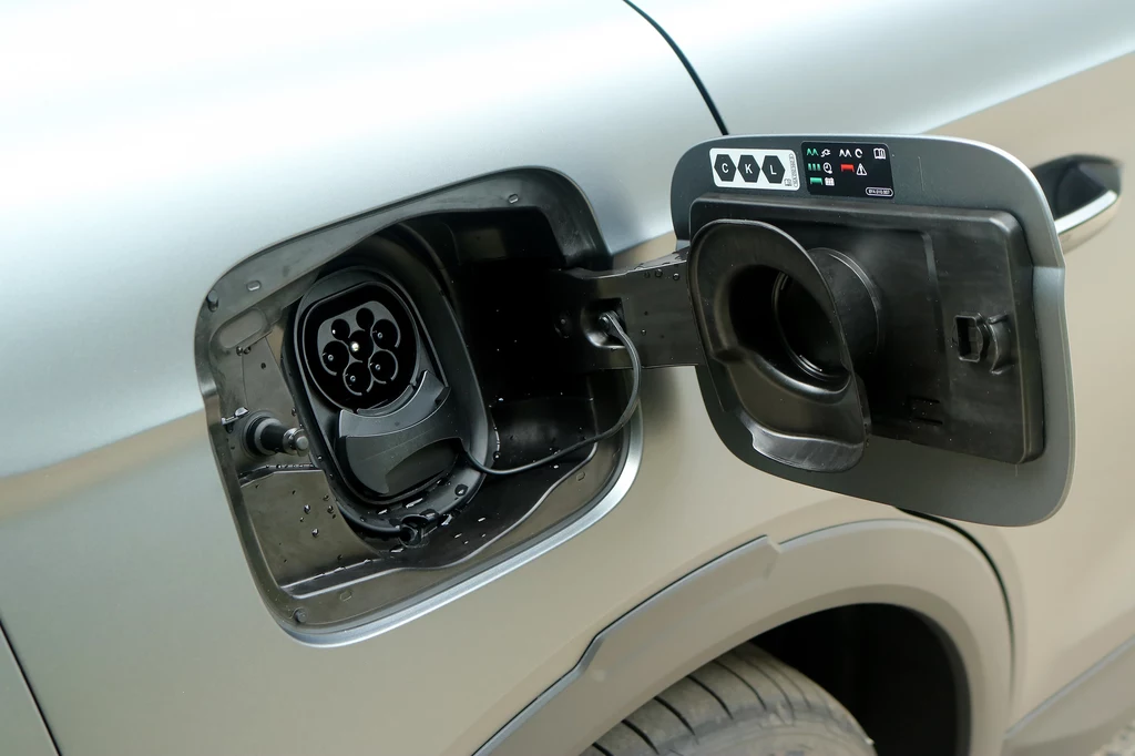 Energię uzupełnimi dzięki ładowaniu o mocy sięgającej 185 kW, co pozwala na naładowanie samochodu do 80 proc. w zaledwie 26 minut. Wlot gniazda ładowania samochodu elektrycznego widoczny po otwarciu klapki, obok otworu ukryte elementy panelu kontrolnego; karoseria w kolorze srebrnym, fragment koła w tle.