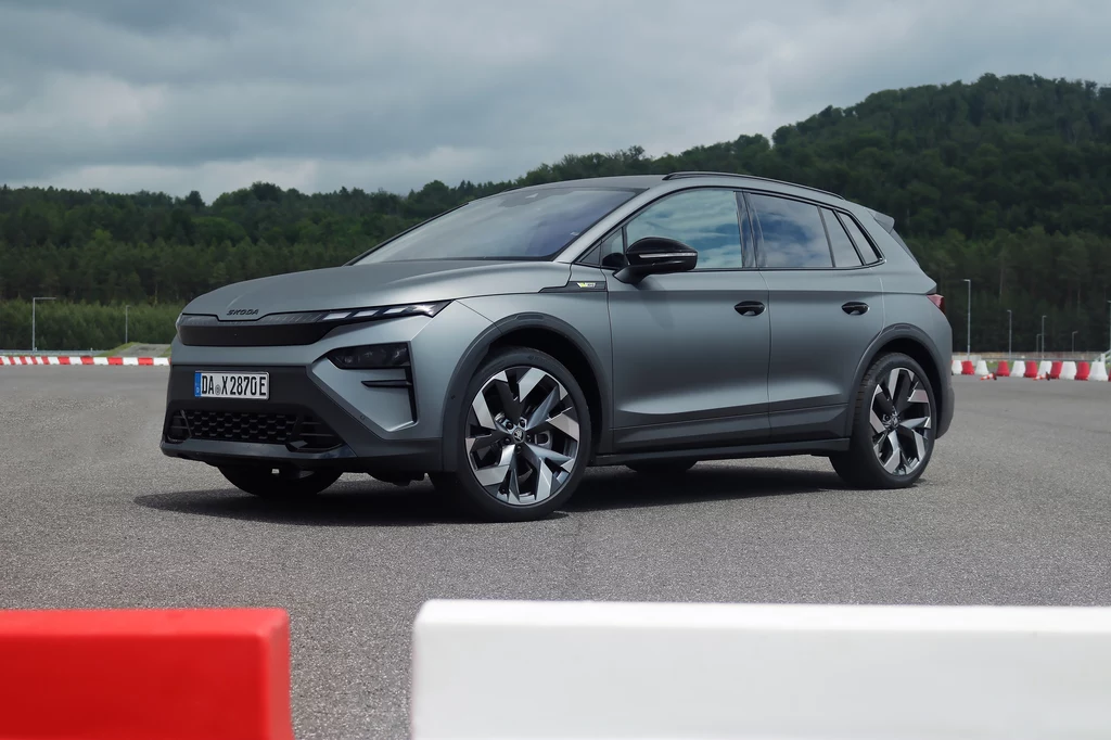 Adaptacyjne zawieszenie DCC pozwala na dynamiczną jazdę nawet po torze. Nowoczesny elektryczny SUV w matowym szarym kolorze stoi na torze testowym z barierami, w tle widoczne zielone wzgórza oraz pochmurne niebo.