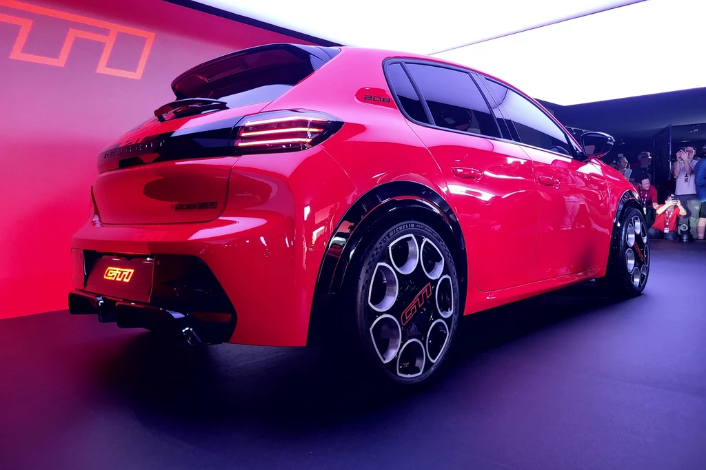 Czerwony lakier nawiązuje do koloru protoplasty z lat 80., czyli modelu 205 GTi Czerwony samochód osobowy w wersji sportowej ustawiony na ekspozycji, podświetlony dynamicznym światłem, z widocznym logo GTI na klapie bagażnika i felgach, wokół publiczność obserwująca prezentację.
