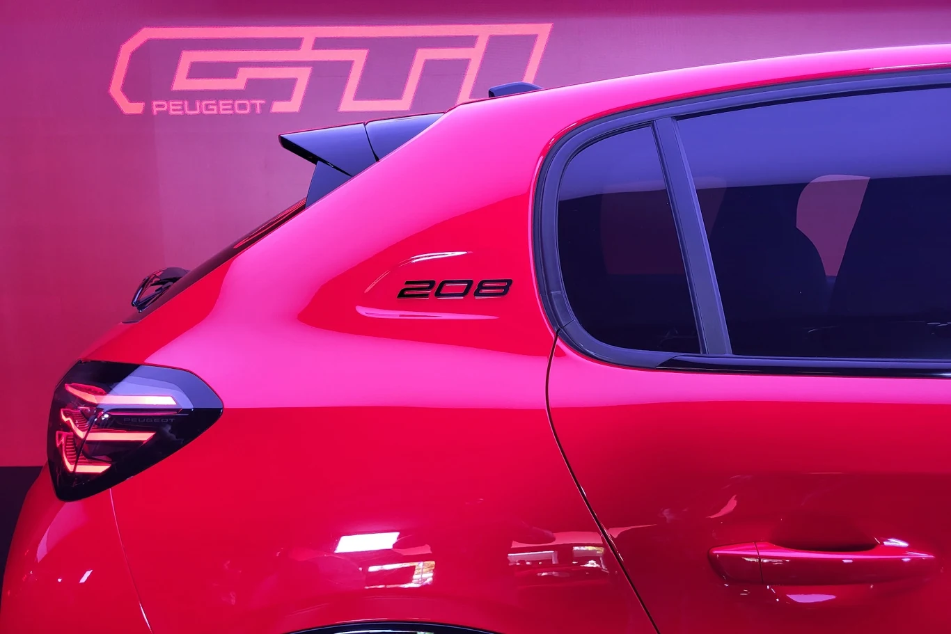 Tylna część czerwonego samochodu Peugeot 208, widoczne światło tylne, przyciemniane szyby oraz oznaczenie modelu na karoserii, w tle neonowy napis GTI.