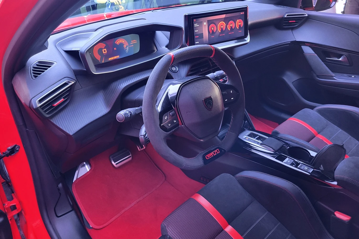 Wnętrze nowoczesnego sportowego samochodu z cyfrowym zestawem wskaźników, dużym ekranem na kokpicie oraz kierownicą pokrytą alcantarą z napisem GTI, fotele z czarno-czerwonym wykończeniem.