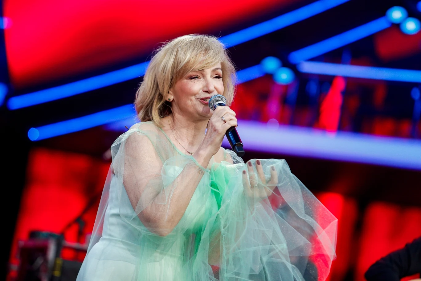 Kobieta o blond włosach śpiewa na scenie, ubrana w jasnozieloną kreację z tiulu, trzyma mikrofon, w tle kolorowe światła i rozmyte elementy scenografii.