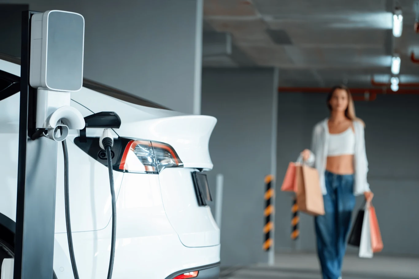 Zainteresowanie programem rządowym "NaszEauto" rośnie. Wypłacono 63 wnioski. Biały samochód elektryczny podłączony do stacji ładowania w podziemnym garażu, w tle kobieta trzymająca torby zakupowe zbliża się w kierunku pojazdu.