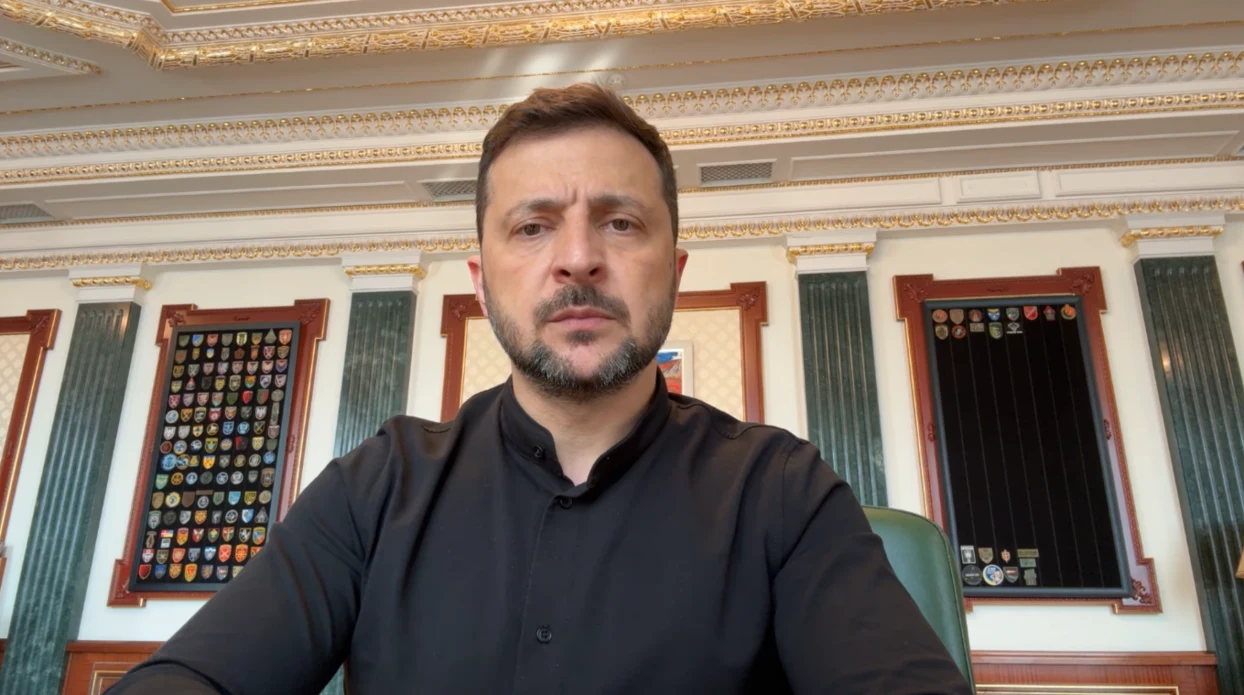Wołodymyr Zełenski o konflikcie zbrojnym Izraela z Iranem Mężczyzna w ciemnej koszuli siedzi przy biurku w eleganckim wnętrzu, za nim na ścianie widoczne są dekoracyjne elementy oraz tablice z odznakami.