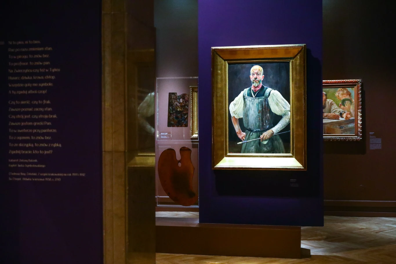 Wnętrze galerii sztuki z dziełem malarskim w złotej ramie przedstawiającym mężczyznę w tradycyjnym stroju, na głównym planie fioletowa ściana ekspozycyjna, w tle inne obrazy oraz paleta malarska.
