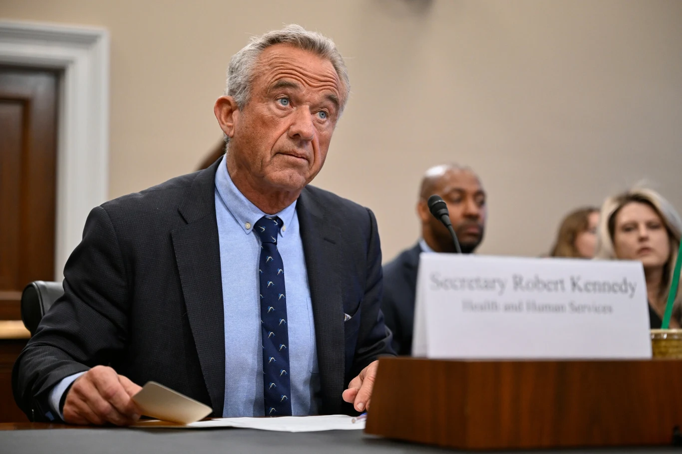 Robert F. Kennedy Jr. powiela teorie spiskowe? Mężczyzna w garniturze siedzący przy stole konferencyjnym, trzymający dokumenty i patrzący w górę, przed nim tabliczka z imieniem i nazwiskiem. W tle widoczne inne osoby uczestniczące w spotkaniu.