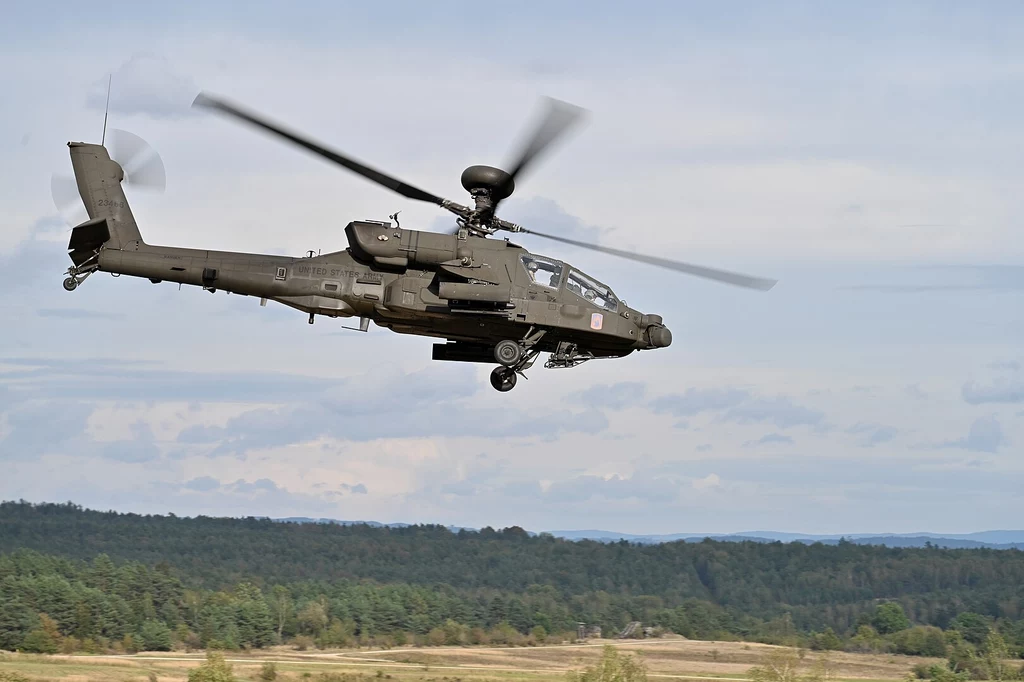 Smigłowiec AH-64E Apache Śmigłowiec wojskowy unoszący się nad rozległym terenem zalesionym, w tle niebo z lekkim zachmurzeniem i horyzont leśny.
