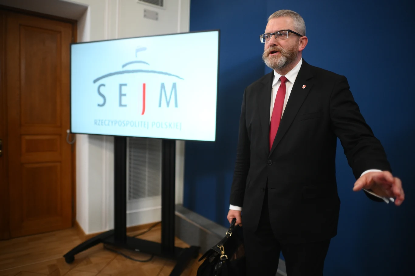 Grzegorz Braun zniszczył wystawę w Sejmie. Lewica składa zawiadomienie Mężczyzna w garniturze i czerwonym krawacie stoi przy ekranie z napisem Sejm Rzeczypospolitej Polskiej, trzymając w ręku torbę, w tle jasna ściana oraz drzwi.