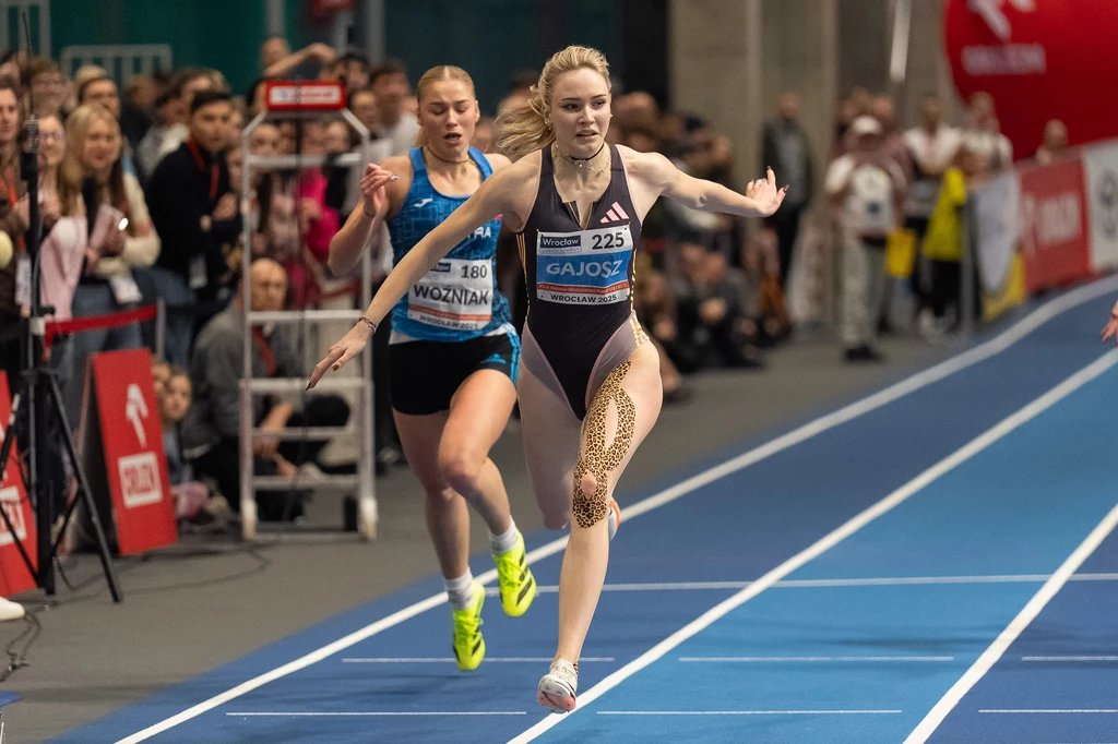 Wiktoria Gajosz Finisz biegu lekkoatletycznego na hali, dwie zawodniczki w sportowych strojach walczą o zwycięstwo na oczach zgromadzonej publiczności, jedna z nich wyraźnie wysuwa się na prowadzenie z wyciągniętymi ramionami blisko mety.