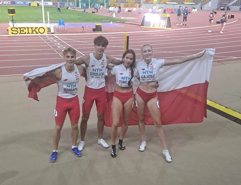 Wiktoria Gajosz (z prawej) wicemistrzynią świata U-20 w sztafecie mieszanej 4x400 metrów (Lima 2024) Czworo młodych lekkoatletów w sportowych strojach reprezentacyjnych pozuje razem na bieżni stadionu, trzymając rozpostartą biało-czerwoną flagę Polski, w tle widoczny fragment trybun i torów lekkoatletycznych.
