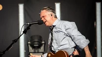 Mężczyzna z gitarą elektryczną gra i śpiewa do mikrofonu na scenie koncertowej, ubrany w rozpiętą koszulę, wyrażając duże emocje podczas występu, w tle widoczne pionowe oświetlenie estradowe.