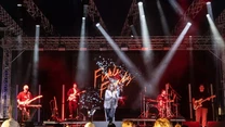 Grupa muzyków występuje na oświetlonej scenie, w centrum wokalistka, a w tle napis z nazwą zespołu; na pierwszym planie publiczność oglądająca występ.
