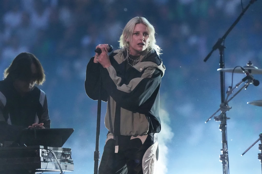 Linkin Park Piosenkarka z jasnymi włosami trzyma mikrofon i występuje na scenie przy świetle reflektorów, obok niej znajduje się osoba obsługująca sprzęt muzyczny, w tle widownia i rozmyte światła.